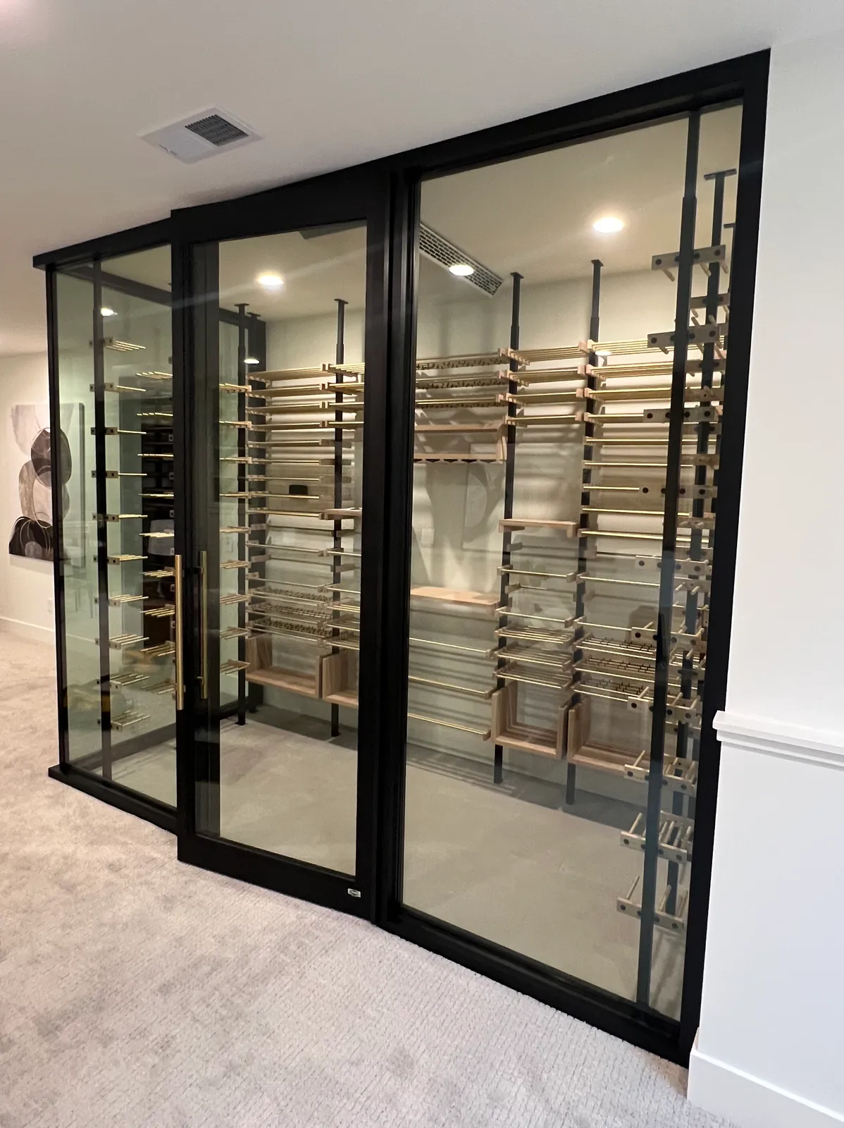Black-framed glass wine room with a full-height glass door and gold pull, wine racks visible inside.