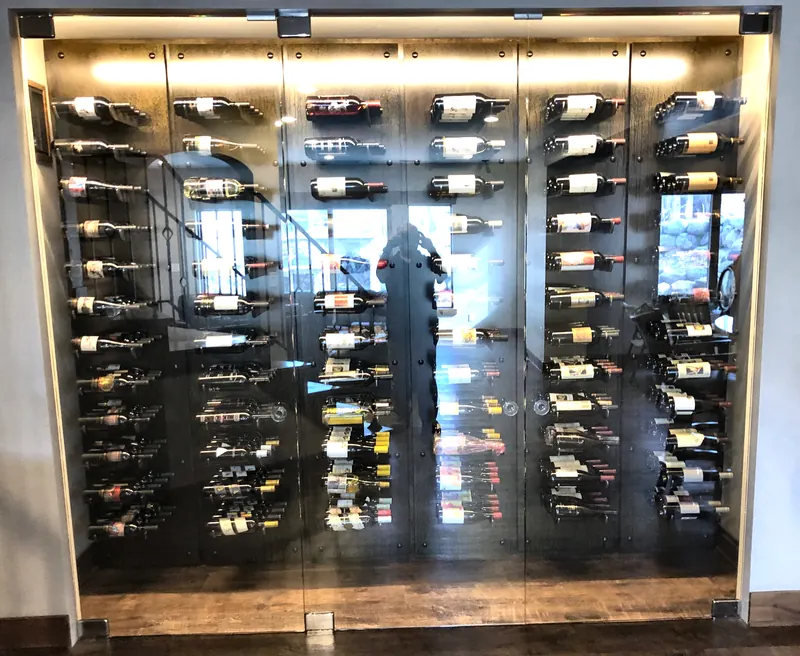Frameless glass wine wall with clamp hardware in front of a lit wall of label-forward bottles.