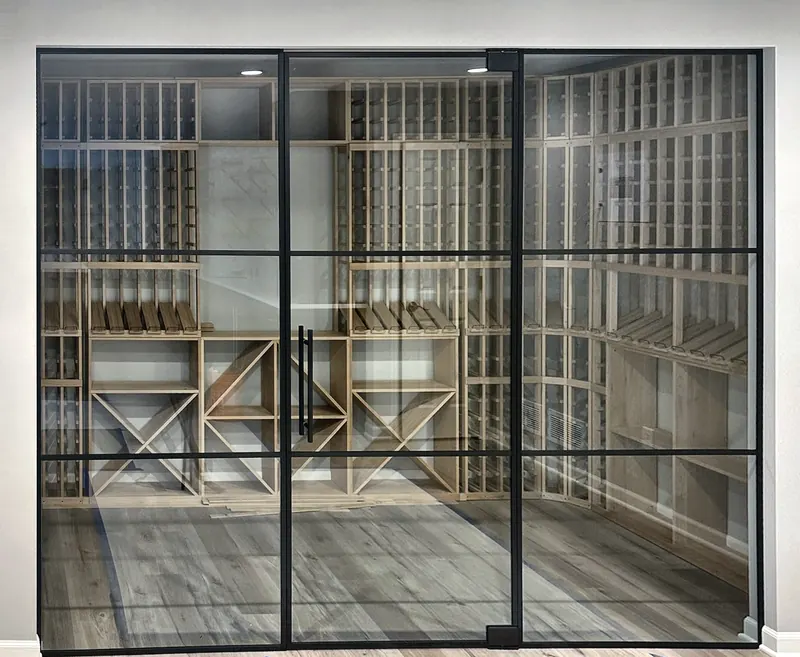 Floor-to-ceiling glass wine room front with a black grid frame and center glass door, wood racking inside.
